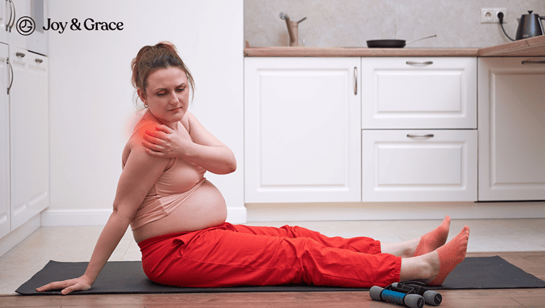 A pregnant woman is experiencing shoulder pain while sitting on a yoga mat in the kitchen.