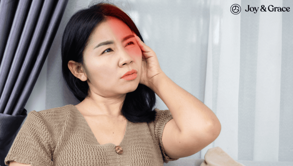 A woman is holding her hand to her head in pain, specifically on the left side of her neck, while sitting on a couch.