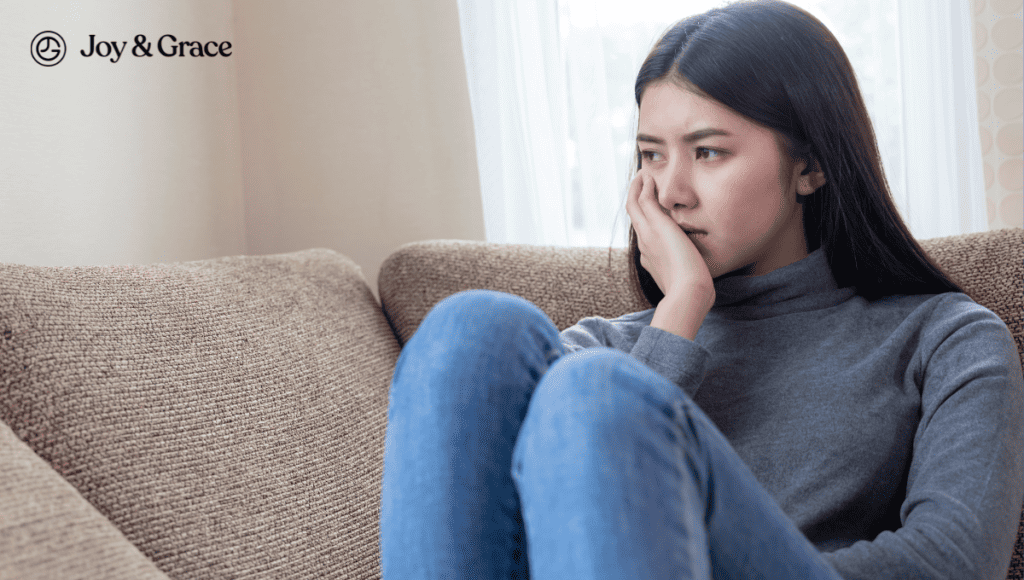 A woman sitting on a couch with her hand on her left shoulder in pain.