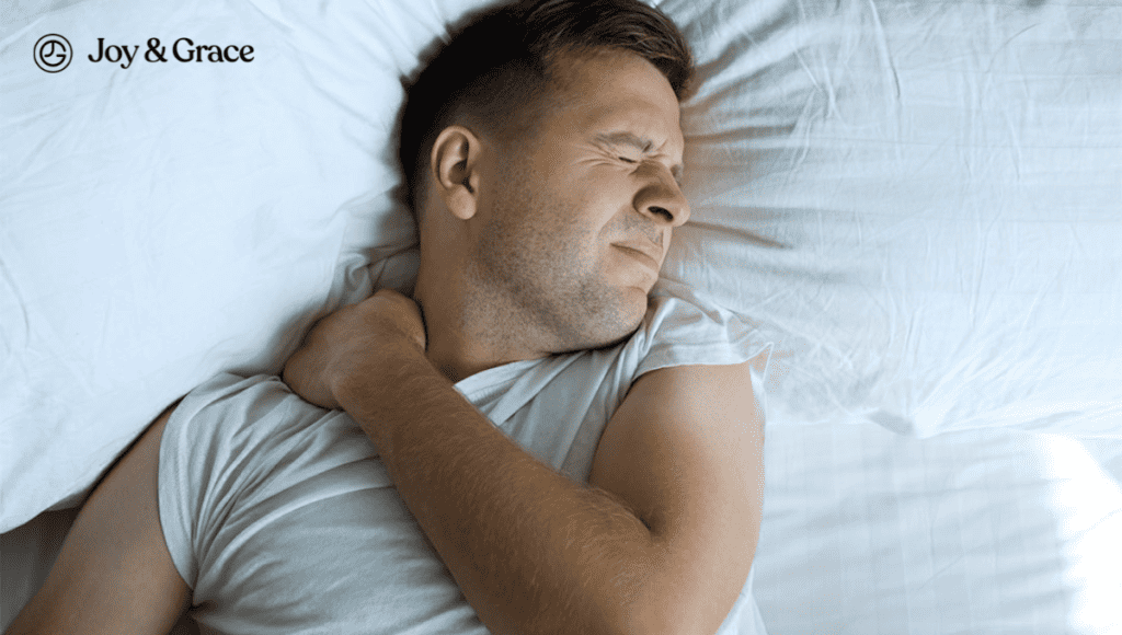 A man is laying in bed in a sleeping position with his hand on his neck, experiencing shoulder blade pain.